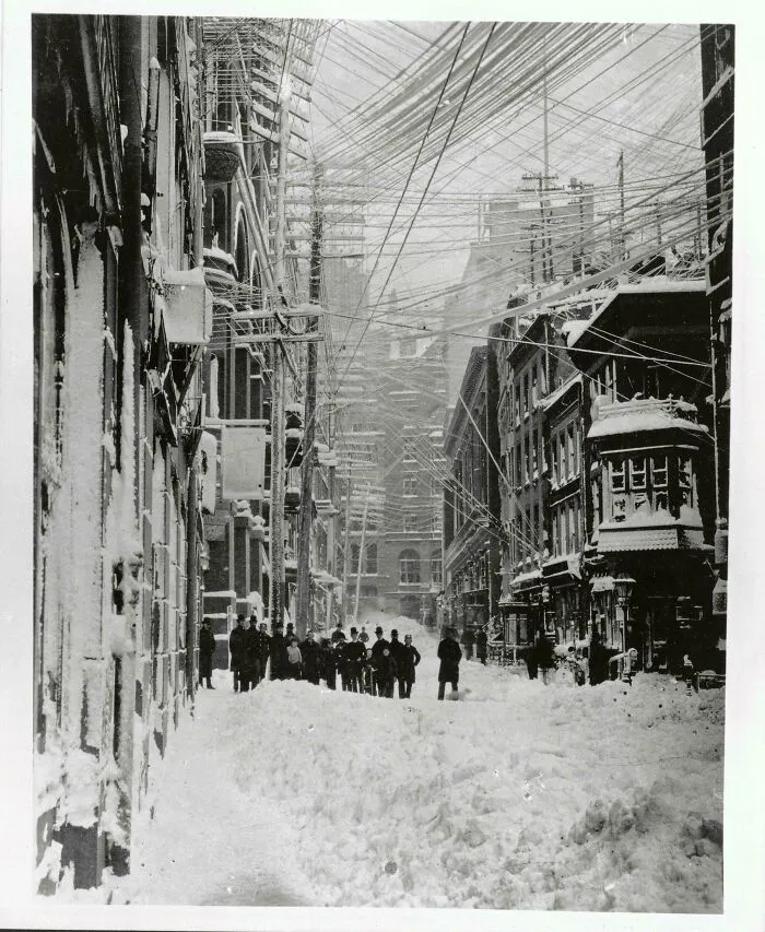 A historic black and white photo showing a snowy city street with numerous power lines overhead.