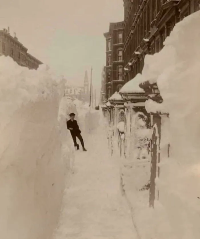 A man stands dwarfed by towering snowdrifts, a fascinating photo from American history during a massive blizzard.