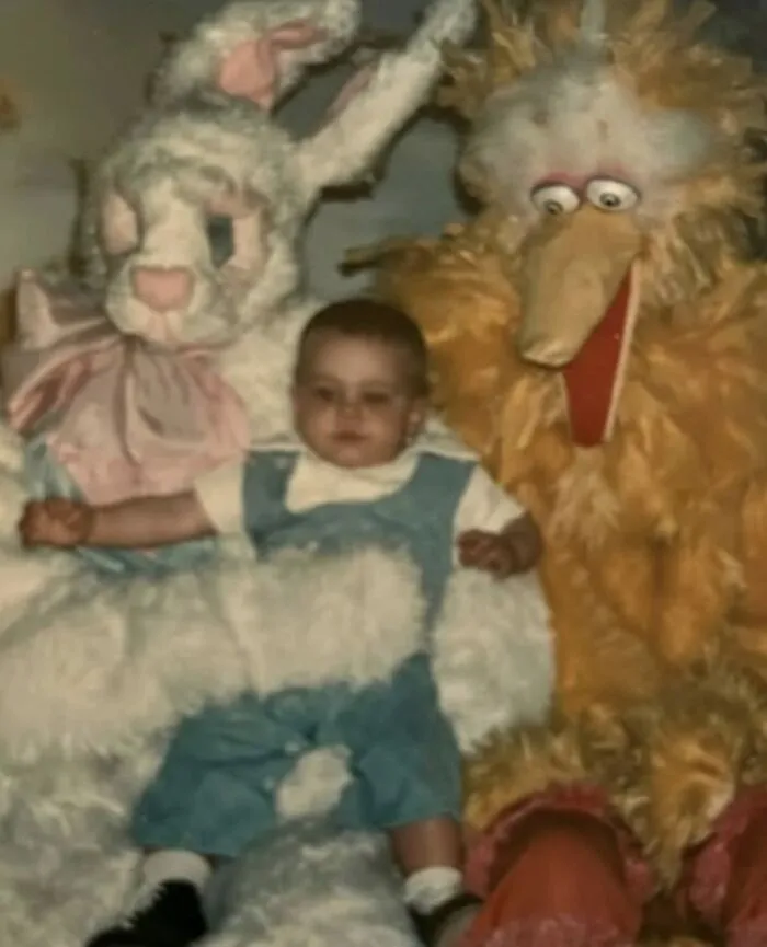 An infant sits uncomfortably between a large white bunny and a terrifying character mascot of Big Bird in an awkward family photo.