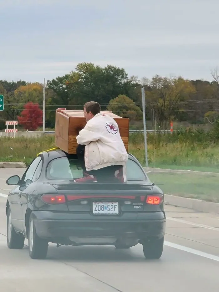Guy Riding Down The Road Holding A Dresser On Top Of A Car On A 50 Mph Road