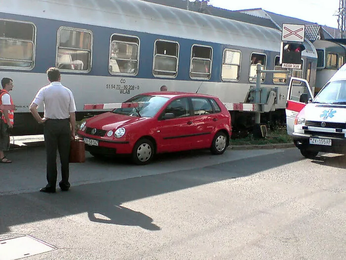 A red car stopped on train tracks with a train behind it and an ambulance, depicting people so dumb.