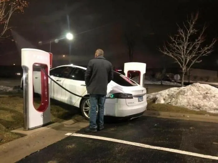 A person standing by a white Tesla plugged into a charger, illustrating dumb positioning.