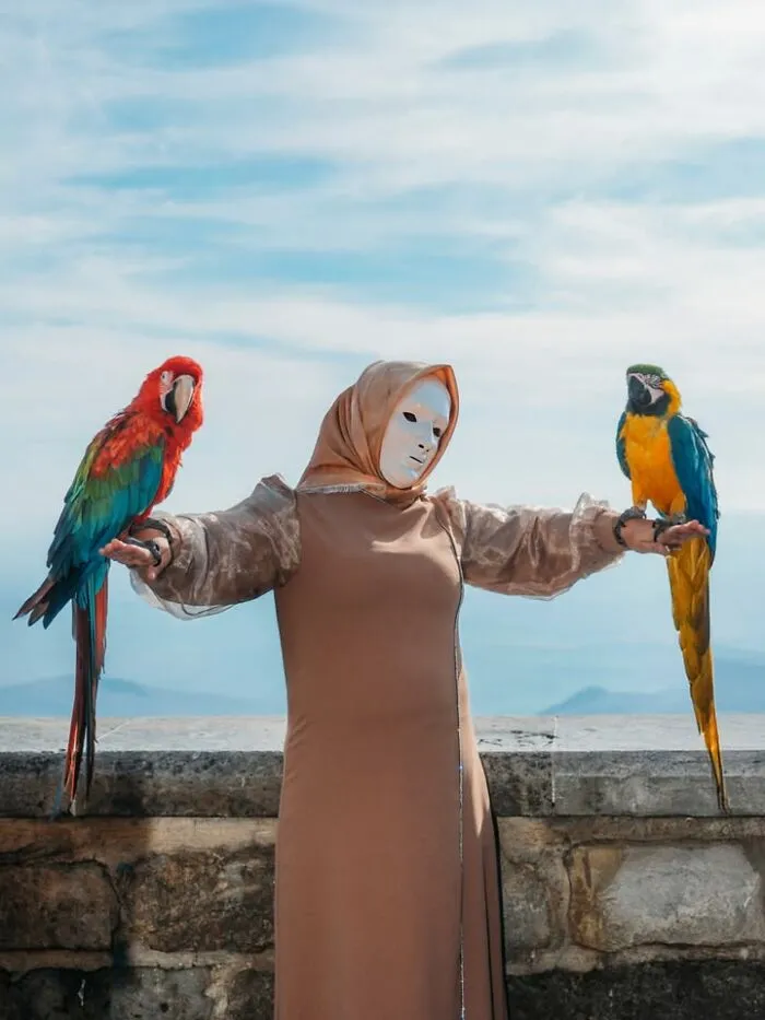 A woman in a mask and long dress with two parrots on her arms, a well-timed street shot.
