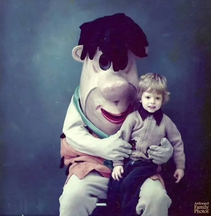 Awkward family photos show a young child sitting calmly with a terrifying character mascot of Fred Flintstone.