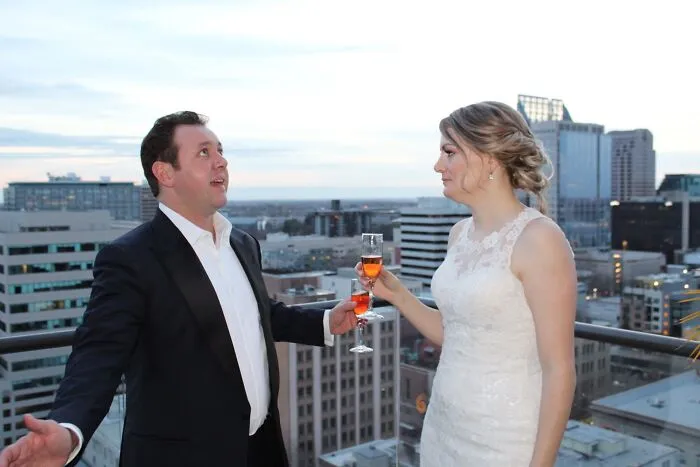 An iconic funny wedding photo of a bride and groom toasting with champagne on a rooftop overlooking a city.