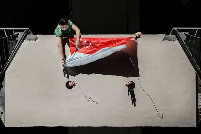 A person holding a Singapore flag while two hands emerge from holes in a wall, for a street photography award.