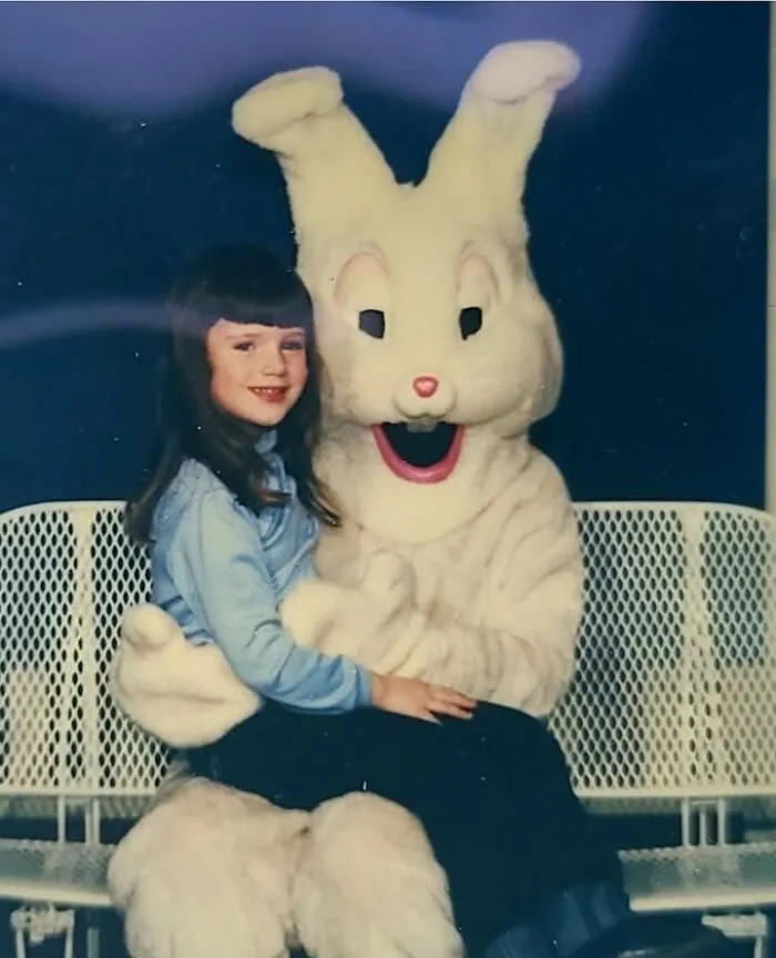 A smiling girl sits on the lap of a slightly terrifying Easter bunny character mascot for an awkward family photo.