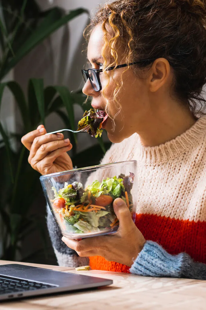 A woman eats a healthy salad while looking at a laptop. Eating healthy foods is one of the important healthy habits.