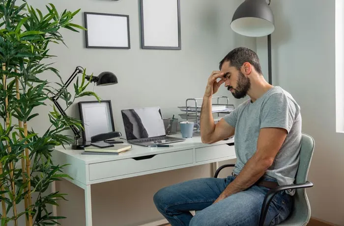 A man sits at a desk, head in hand, looking stressed, illustrating the impact of dumb work rules.