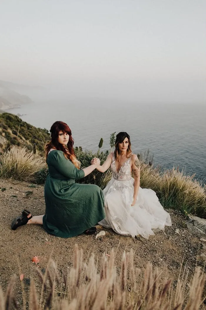 Two women pose on a cliff overlooking the ocean, an iconic and funny wedding photo.
