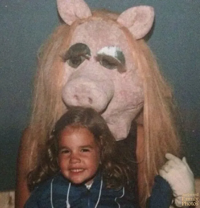 Awkward family photos: A smiling child sits in front of a truly terrifying pig character mascot with long hair.