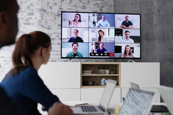 Two employees look at a screen displaying a video conference with multiple people, illustrating dumb work rules.