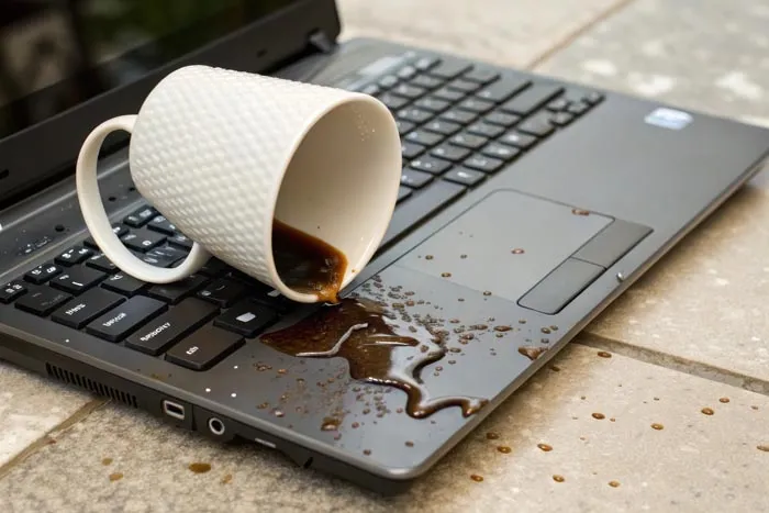 A white mug spilling coffee onto a laptop keyboard, representing dumb work rules.