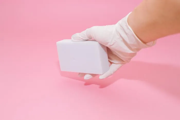 A gloved hand holding a white magic eraser against a pink background, showcasing dumb work rules.