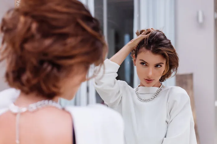 A woman with short hair looking at her reflection in a mirror, adjusting her hair, considering healthy habits.