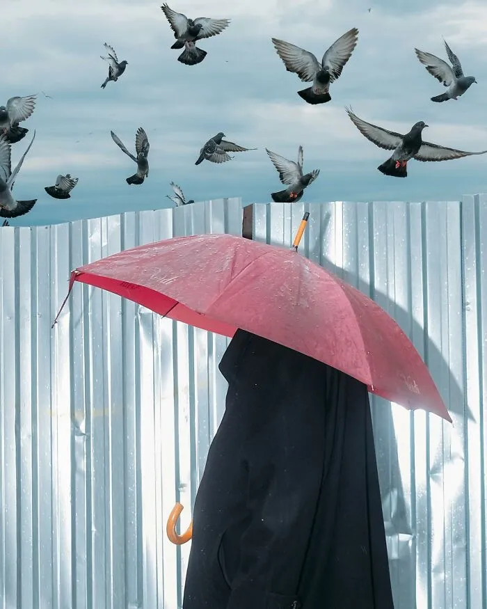 A photographer captured a ridiculously well-timed street shot of a red umbrella and many pigeons flying in the sky.