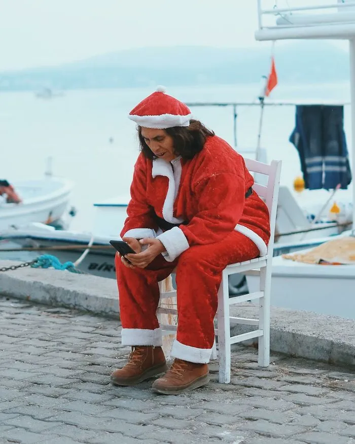 A person dressed as Santa Claus checking their phone while sitting on a chair by the water, a well-timed street shot.