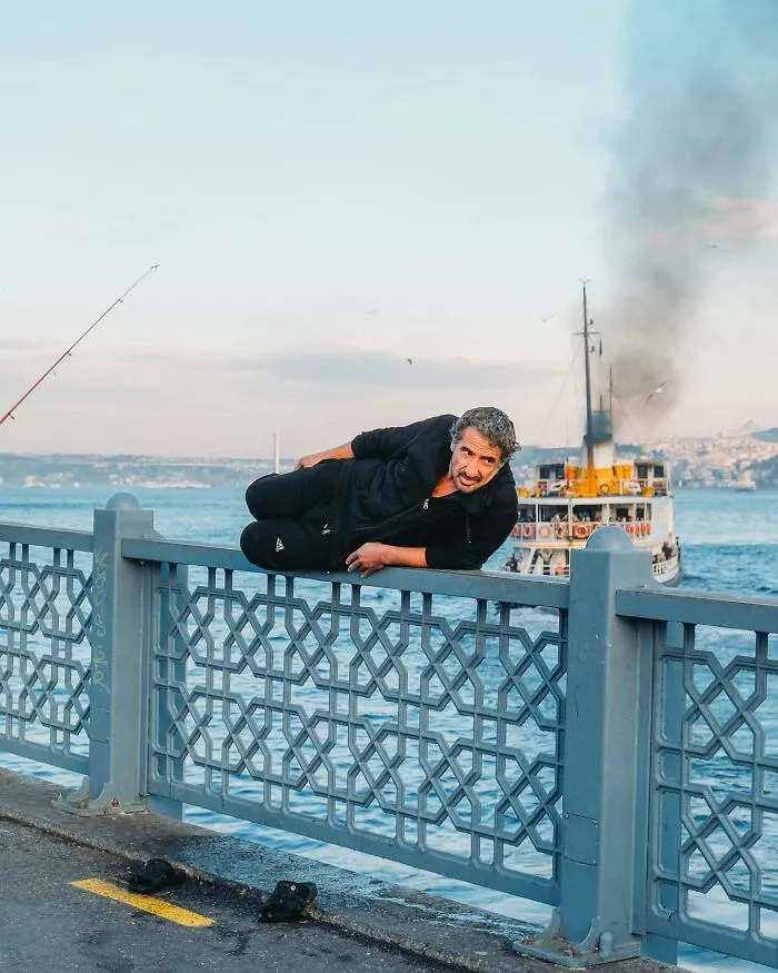 A photographer captured a ridiculously well-timed street shot of a man lying on a bridge railing with a boat in the background.