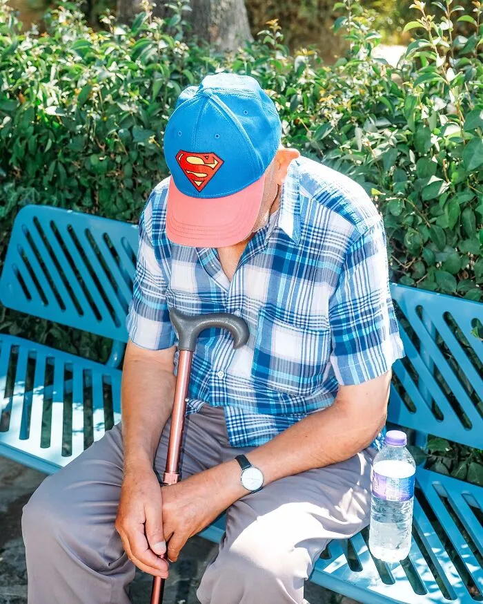 An elderly man wearing a Superman hat sits on a bench with a cane, a well-timed street shot.