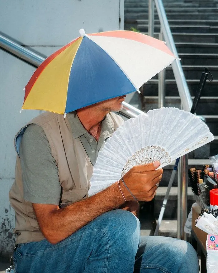 A man with a small umbrella hat and a white fan, captured in a well-timed street shot.