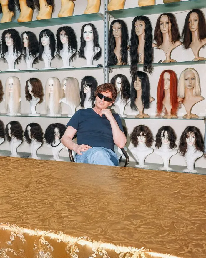 A man in sunglasses sits among mannequins with various wigs, a well-timed street shot.