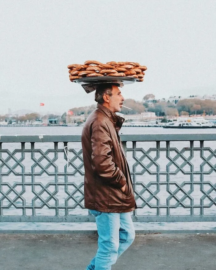 A photographer captured well-timed street shots of a man balancing a tray of bread on his head, walking by water.