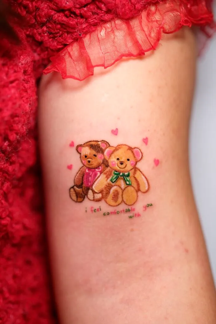 Two adorable teddy bear tattoos with hearts and a quote, resembling childhood crayon drawings, on an arm.