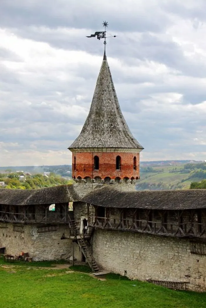 An ancient stone castle tower with character, featuring a conical roof and a weathered brick upper section under a cloudy sky.