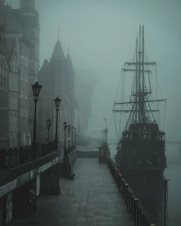 A misty harbor scene with an old sailing ship docked next to historic buildings with character.