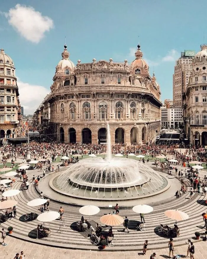 Grand, historic building with character and a large fountain in a bustling city square, surrounded by people and umbrellas.