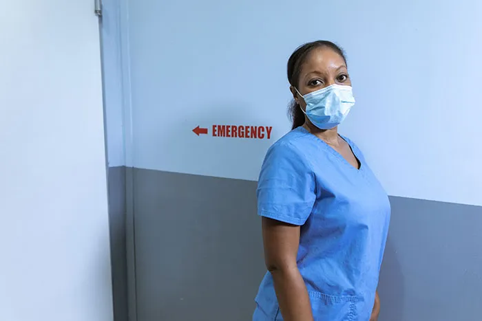 A nurse wearing a mask in a hospital hallway, near an emergency sign, listening to patients saying weird things.