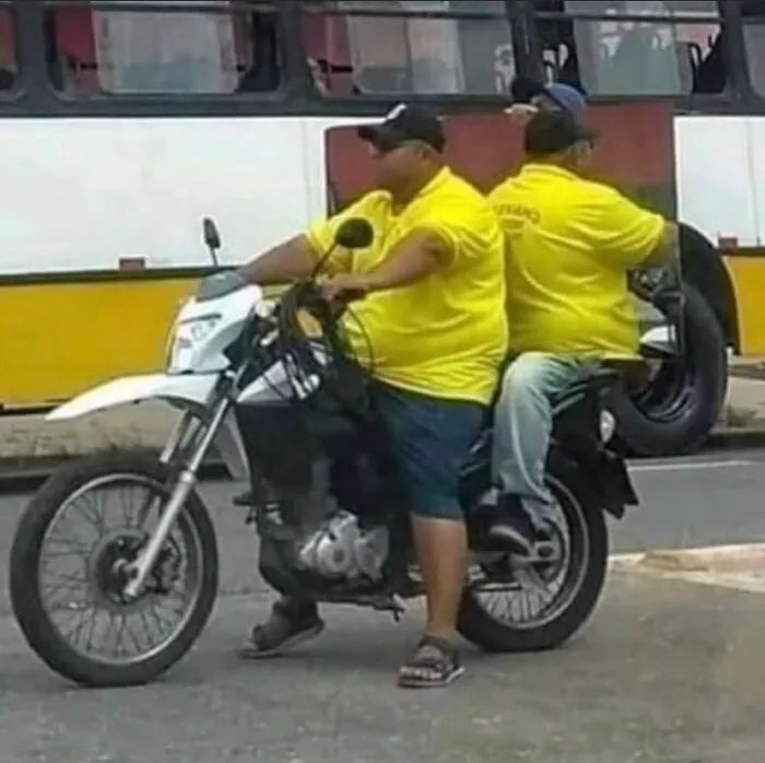 Two men in yellow shirts, sitting on a single motorcycle, with one looking like he is sitting on another man's lap, a cursed image.