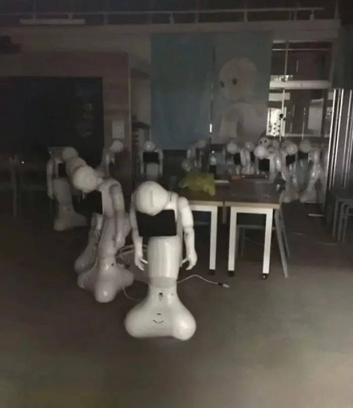 A dark room filled with several white humanoid robots standing idly, an unhinged and cursed image.