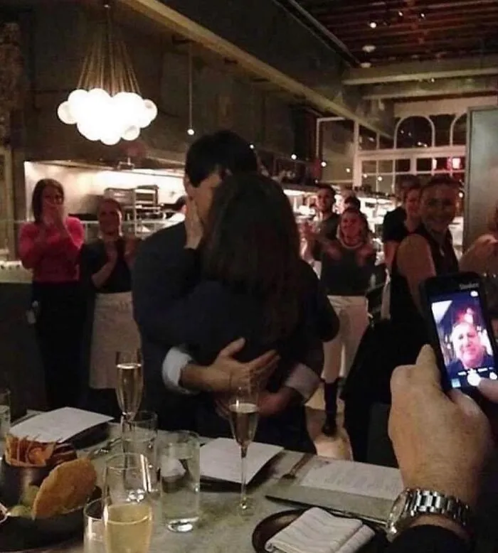 A cursed image of a couple hugging in a restaurant, surrounded by applauding people, while someone takes a photo with a phone.