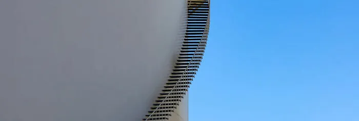 Minimalist photography of a winding staircase on the side of a large white cylindrical structure against a clear blue sky.