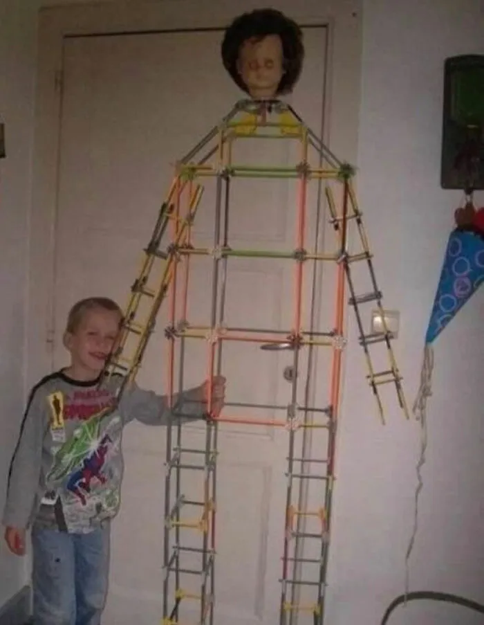 A smiling young boy standing next to a tall, human-like structure made of colorful sticks with a doll head, a random image.