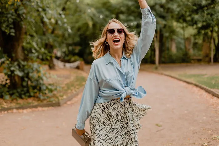 Woman in sunglasses and a denim shirt cheering outdoors, a real deal sign from the universe.