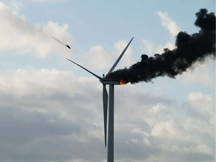 Unsettling photo of a wind turbine on fire with black smoke billowing, a person visible near the flame, and another figure falling.