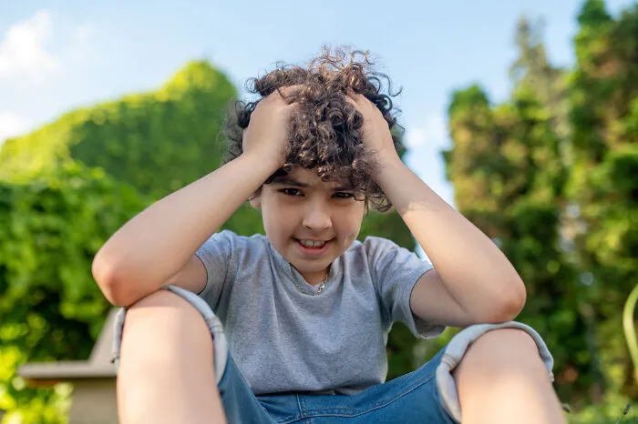 A young boy with curly hair, hands on his head, smiling at the camera, a sign from the universe.