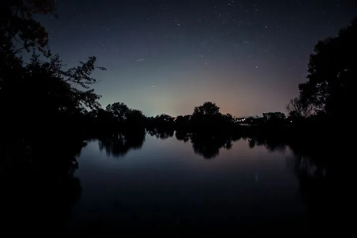 A dark, starry night sky over a tranquil lake with tree silhouettes, illustrating a serene scene for recalling weird encounters.