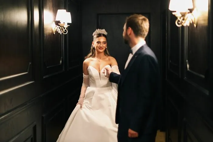 A smiling bride in a white dress and tiara holding hands with a groom, witnessing absurd and hilarious things.