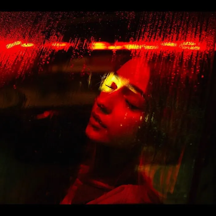 A woman with closed eyes, illuminated by red neon lights through a wet window, creating a cinematic portrait.