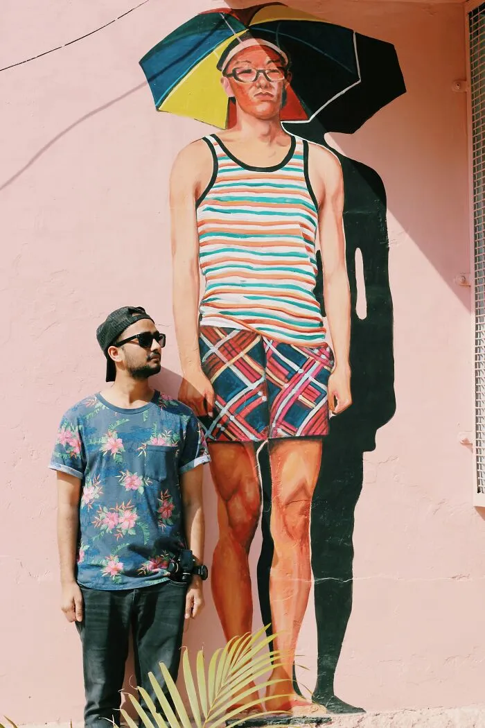 A man in a floral shirt stands next to a mural of a larger man, illustrating gender roles.