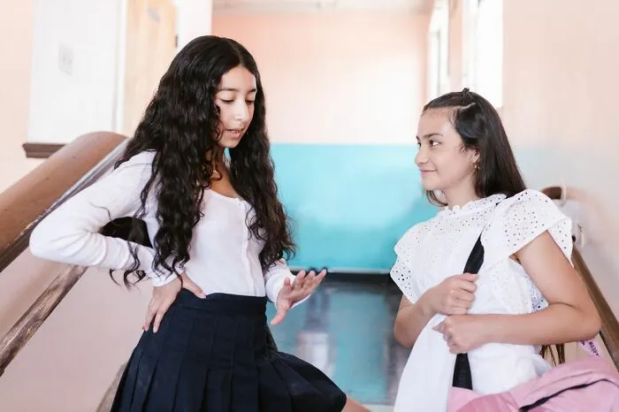 Two girls in school uniforms are talking in a hallway, one with a backpack. This image relates to toxic femininity.