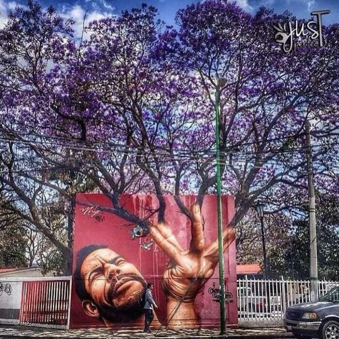 A vibrant street art mural of a man's face and a hand forming a peace sign on a red wall, with purple jacaranda trees above.