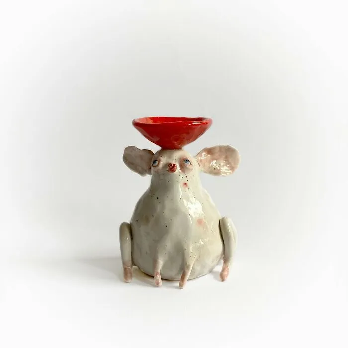 Weird ceramic sculpture of a whimsical white creature holding a small red bowl on its head.