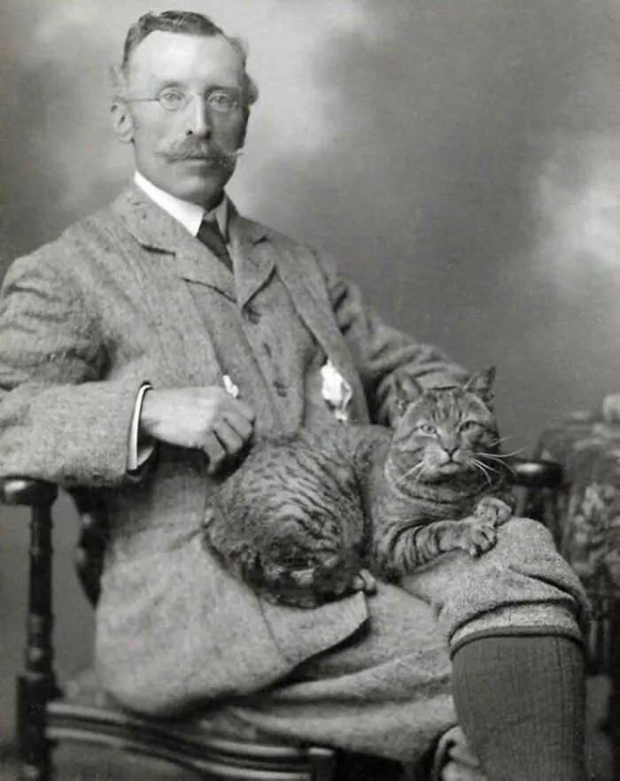 A dapper gentleman posing for a vintage cat photo with a tabby resting on his lap