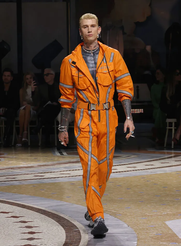 Model wearing bright orange jumpsuit on runway at Plastic Surgery Expo