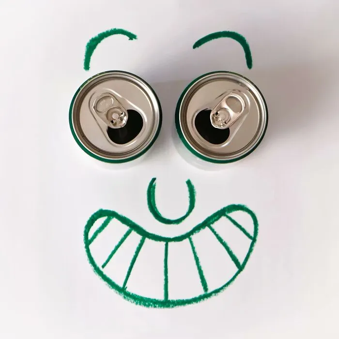Two aluminum cans used as eyes for a drawn smiling face in a playful illustration of everyday objects.