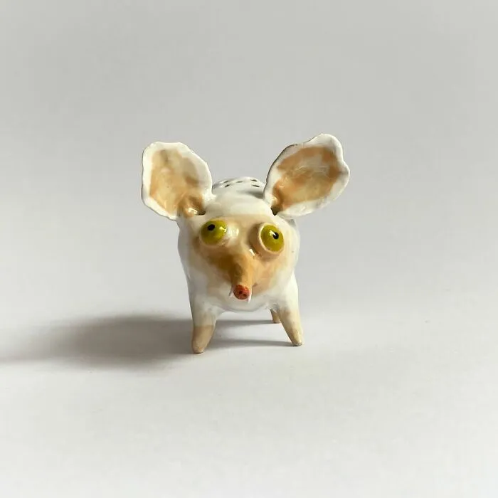 A tiny, quirky ceramic sculpture of a white and yellow creature with big round eyes and ears.